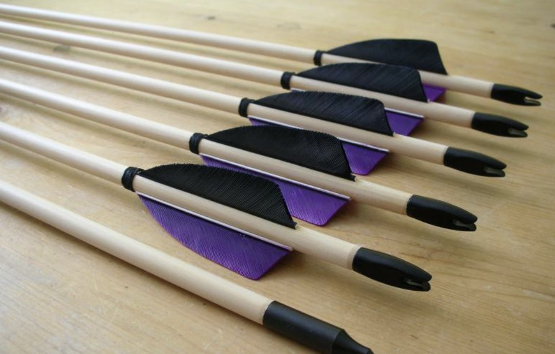 A row of fletched carbon arrow shafts lined up on a wood surface, ready for spine matching