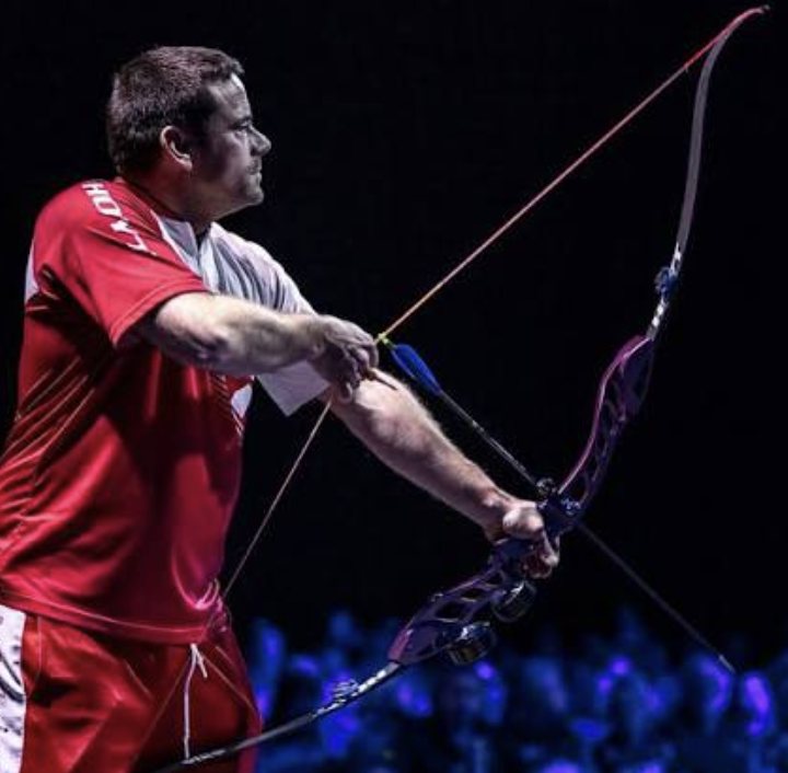 Barebow archer in an indoor arena raising a purple ILF recurve riser with no sight or long rod, three fingers on the string