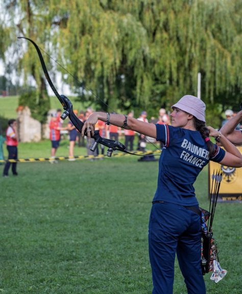 Barebow archer at full draw on an outdoor field, ILF riser with no sight and no stabilizer rods, weight mounted low on the riser