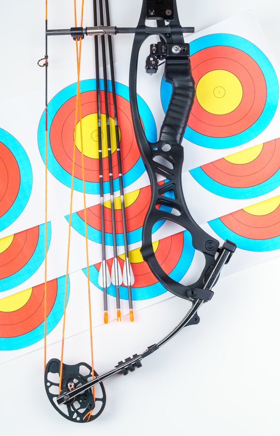 Overhead flat-lay of a compound bow and arrows on a stack of paper target faces