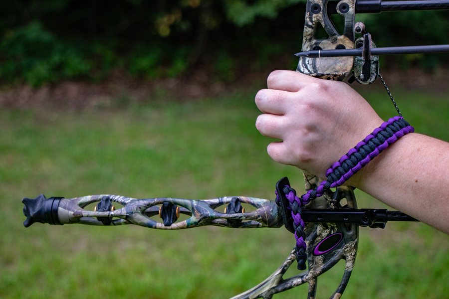 Close-up of an archer's fist wrapped around a camo compound bow grip with a purple paracord wrist sling