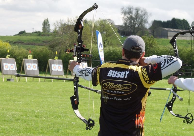 Compound archer at full draw on an outdoor target range, cams and stabilizer visible, with numbered target lanes in the background
