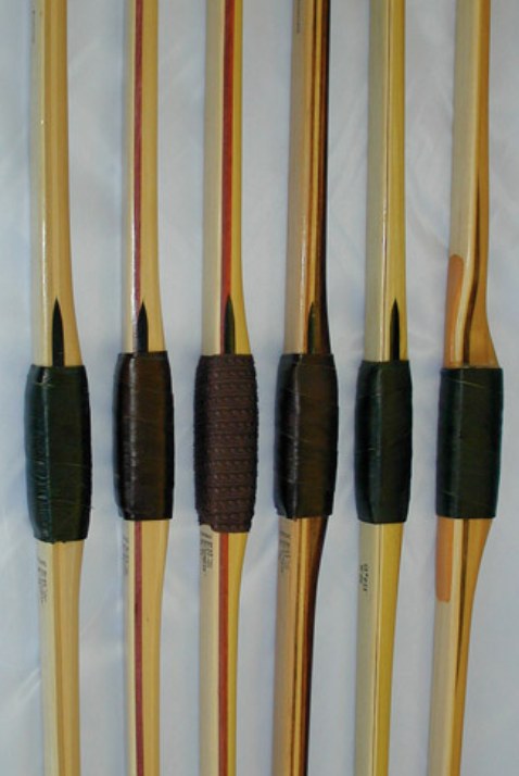 Six wooden bow handles side by side, each with a wrapped grip and a dark tapered arrow-shelf wedge just above it