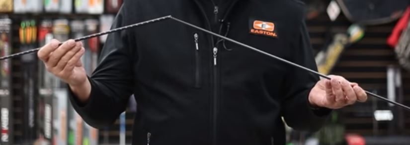 A person in an Easton jacket holding a carbon arrow shaft up between both hands, sighting down its length to check straightness before marking a cutoff point