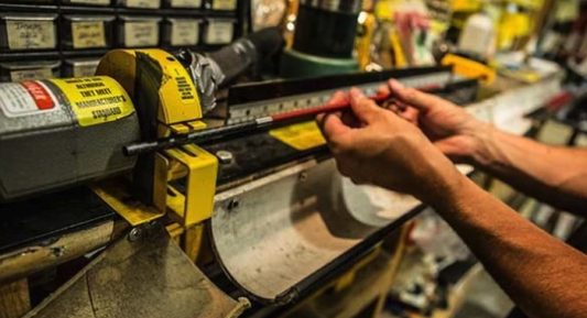 Hands using a marked fixture and pencil to mark the cutoff point on a carbon arrow shaft before cutting