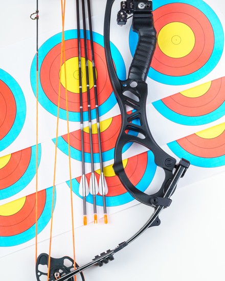 A compound bow riser, cams and three fletched arrows laid across a stack of archery target faces