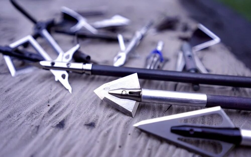 Mechanical and fixed broadheads side by side for comparison An assortment of mechanical and fixed blade broadheads laid out on a workbench next to a carbon arrow shaft for comparison
