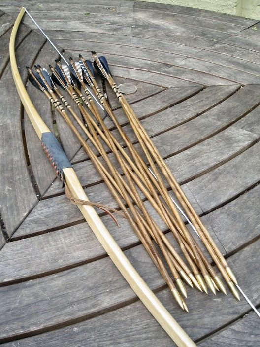 Plain wooden longbow with a leather grip lying on a round slatted table beside a dozen wooden arrows with black and white feather fletching, painted cresting and brass bullet points