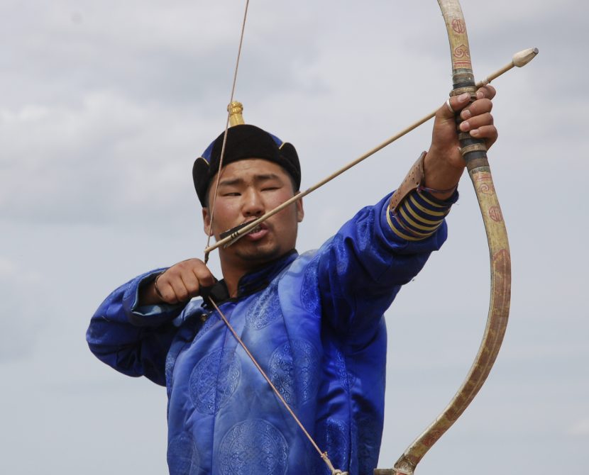 Mongolian archer in a blue silk deel and traditional hat at full draw with a horn composite bow, the string hooked by a closed-fist thumb lock at the cheek and a blunt-tipped arrow resting on the thumb side of the bow