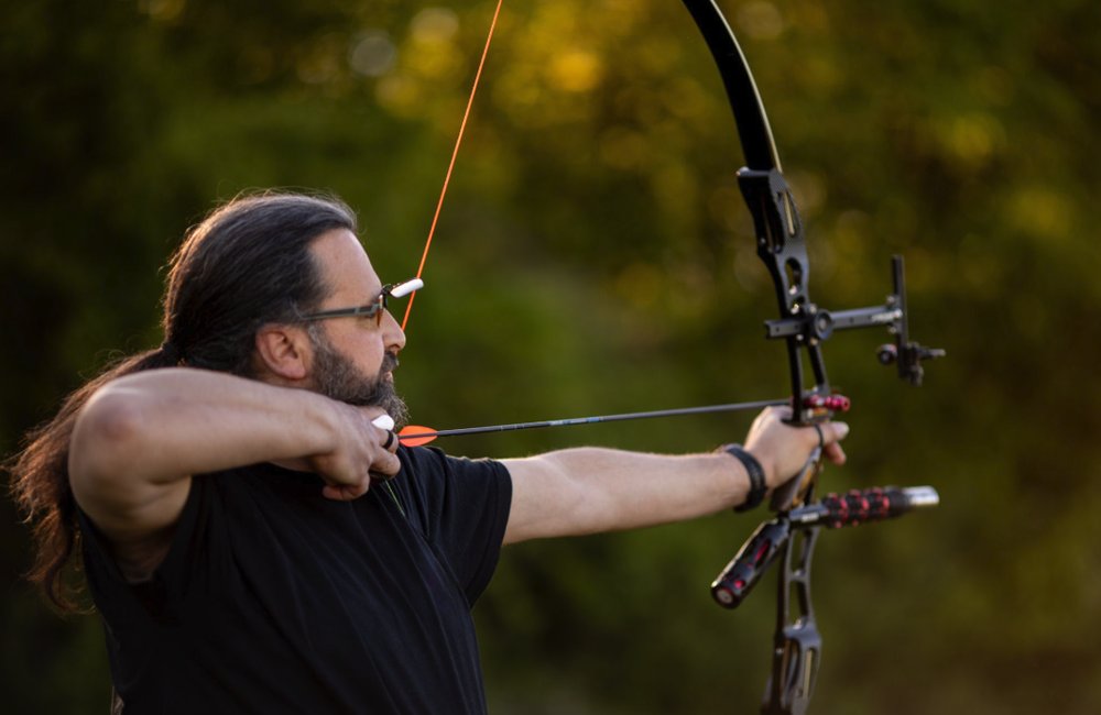 Archer at full draw with a compound bow, cams and sight visible, backlit at sunset