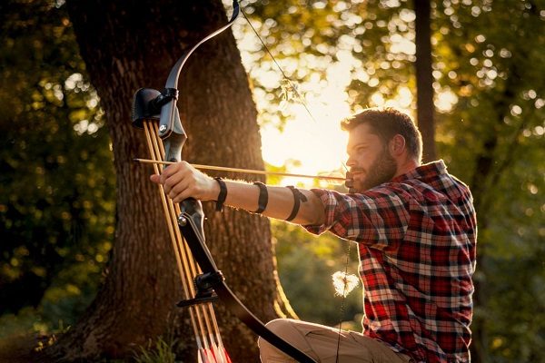 Archer at full draw with a takedown recurve shortbow braced against a tree in wooded terrain