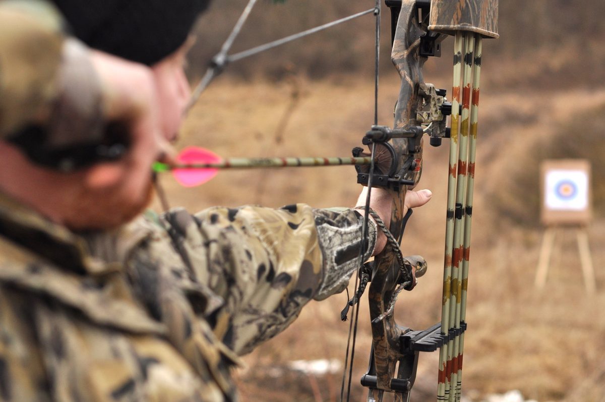 Bowhunter in camo at full draw with a compound bow and a bow quiver of arrows, aiming at a bag target on a wooden stand in an open, dry field