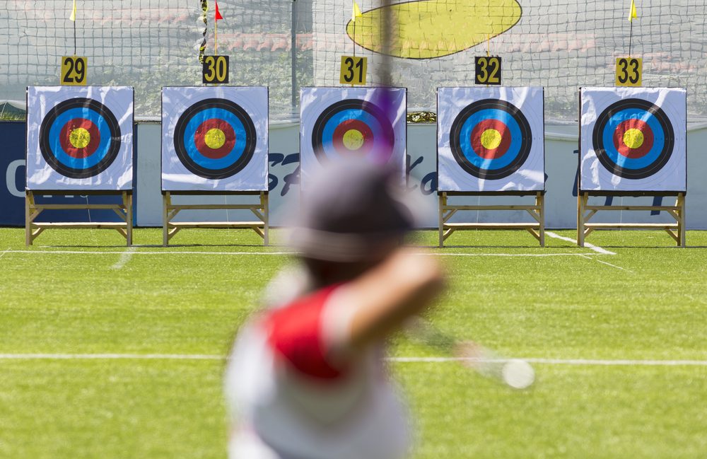 Five target faces on wooden stands numbered 29 to 33 on a mowed field, a white shooting line in front and tall netting behind, with a blurred archer at full draw in the foreground