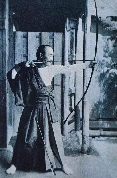 Traditional Yumi archer — full draw form Vintage photograph of a traditional Japanese archer at full draw beside a wooden gate