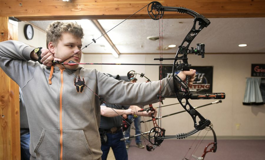 A young man aiming a compound bow at a lit indoor archery range with visible targets downrange