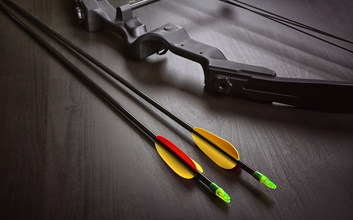 A compound bow with two fletched carbon arrows, one red-and-yellow and one yellow, resting on a dark wood surface