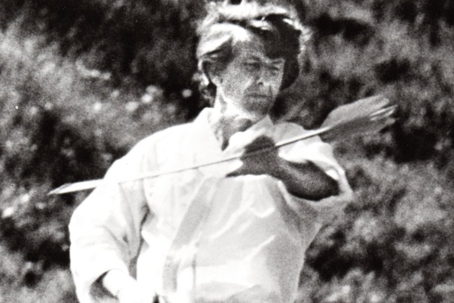 A motion-blurred black and white photograph of an archer at full draw, the loosed arrow already a blur