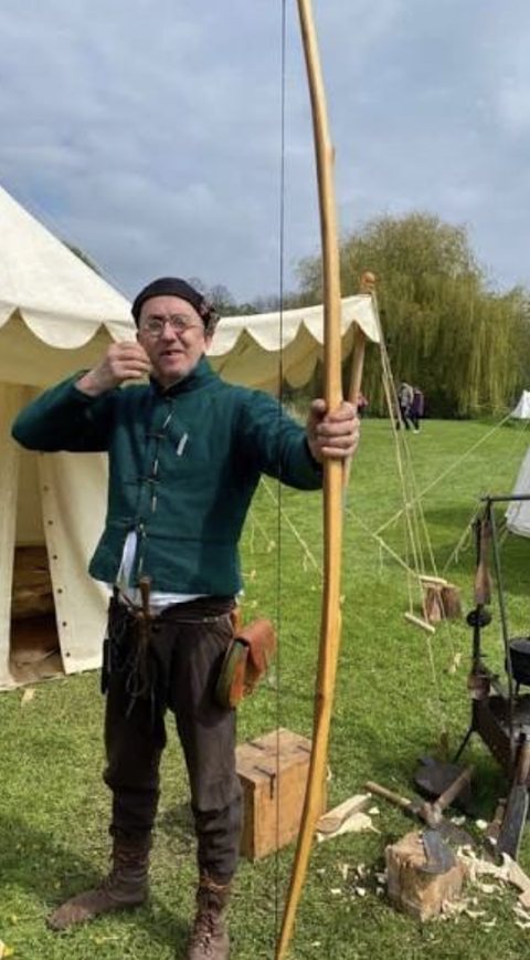 A historical reenactor in period dress drawing a tall wooden English longbow outside a canvas pavilion tent