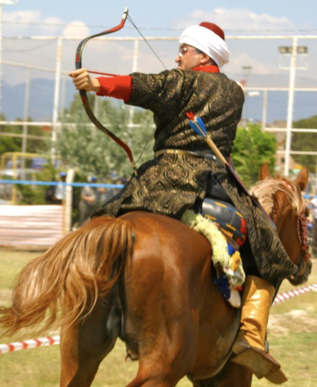 A mounted archer in traditional Ottoman dress drawing a short recurve bow while riding at a gallop