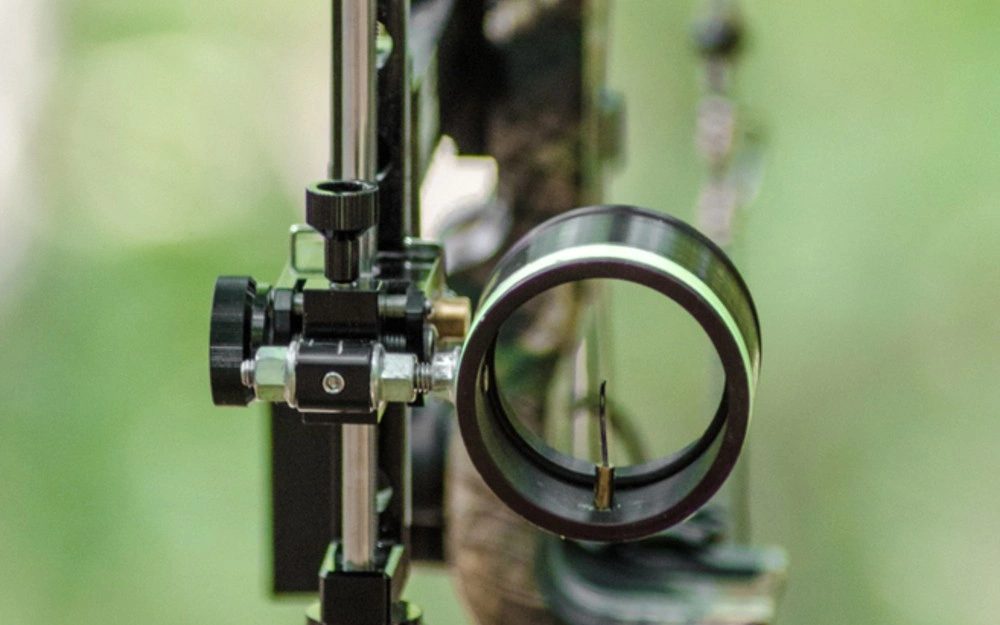 Close-up of a compound bow sight pin and scope housing against green foliage