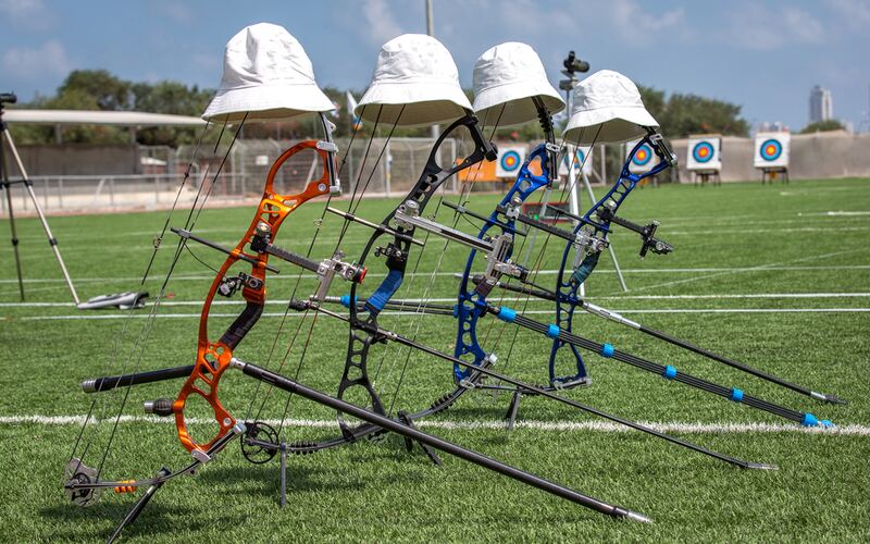 Four recurve and compound bows with white sun hats resting on their sights, lined up on a grass field with target faces in the background