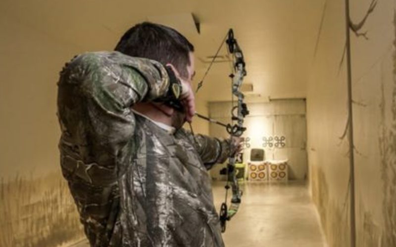 An archer aiming a bow down a narrow indoor corridor toward targets