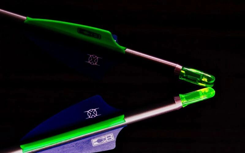Two hunting shafts nock to nock, one green-vaned and one blue-vaned, lit against black