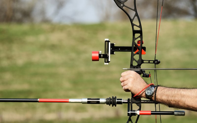 Close-up of a compound bow at full draw, sight and stabilizer visible, on a grass archery field